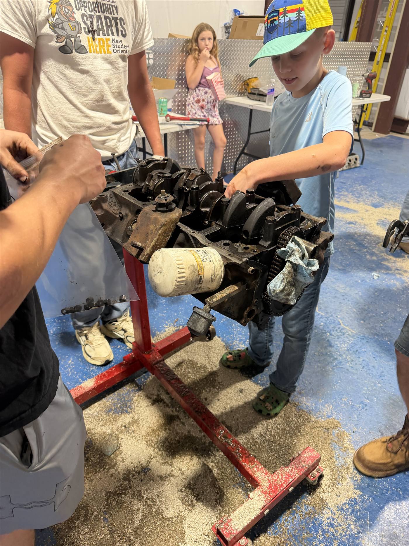 A young student turning the crankshaft of an engine block on a stand while a mentor guides