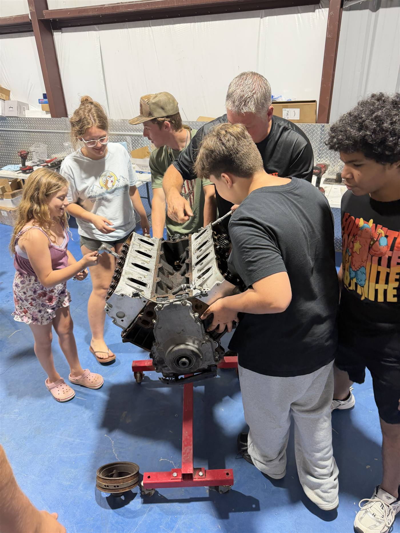 A group of youth and adult mentors gathered around a V8 engine on a stand in the shop, working on it together