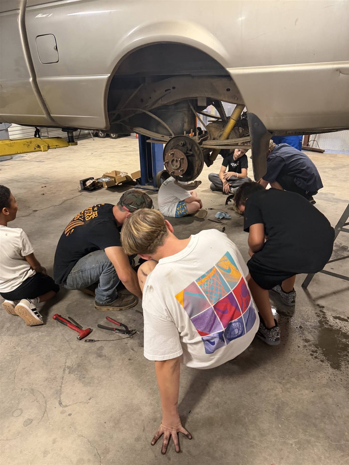 A team of youth crouched under a lifted truck, working together on the rear brakes